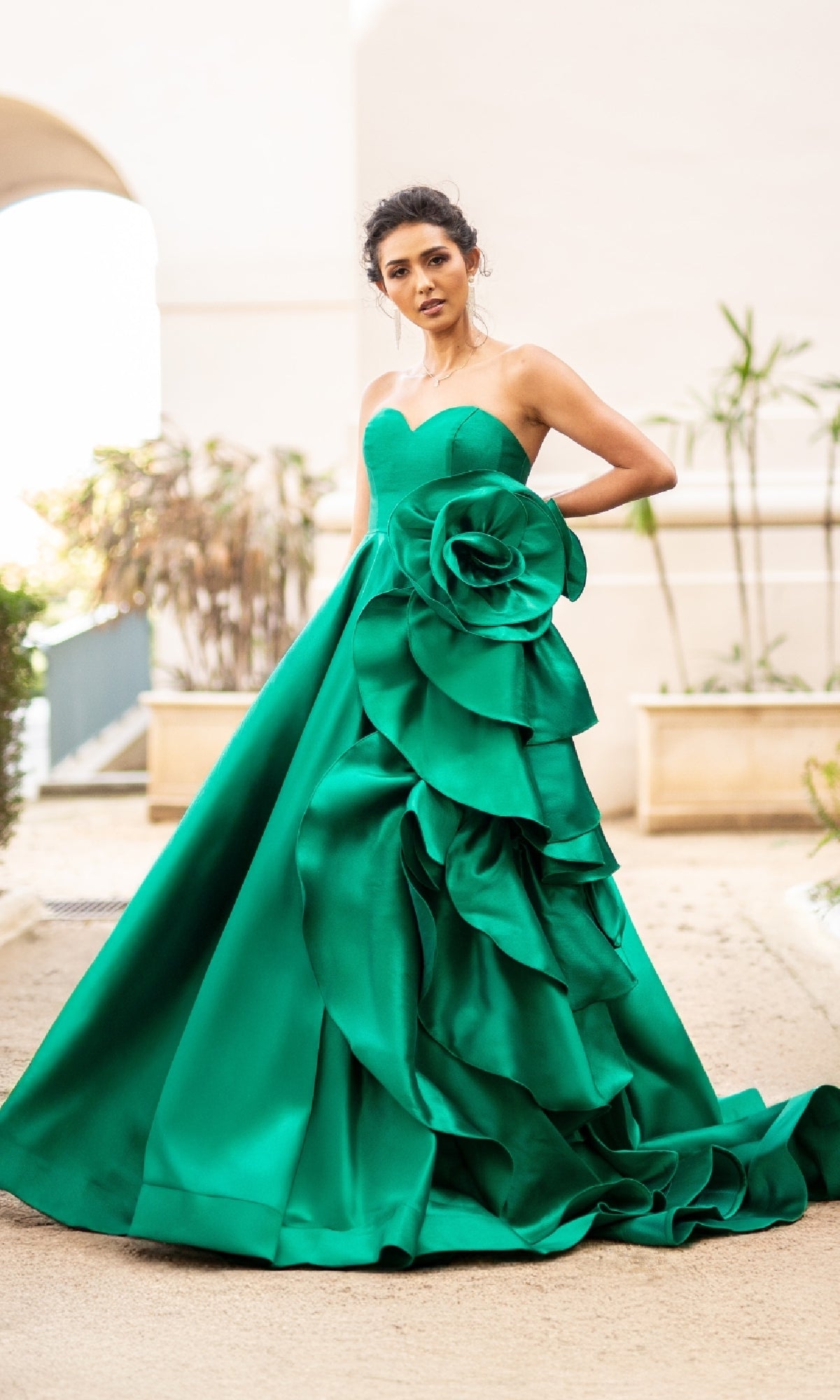 Outdoors, a woman wears the Strapless Prom Ball Gown: Amelia Couture SU085—an emerald green, strapless sweetheart ball gown featuring a large layered rose detail on the skirt.